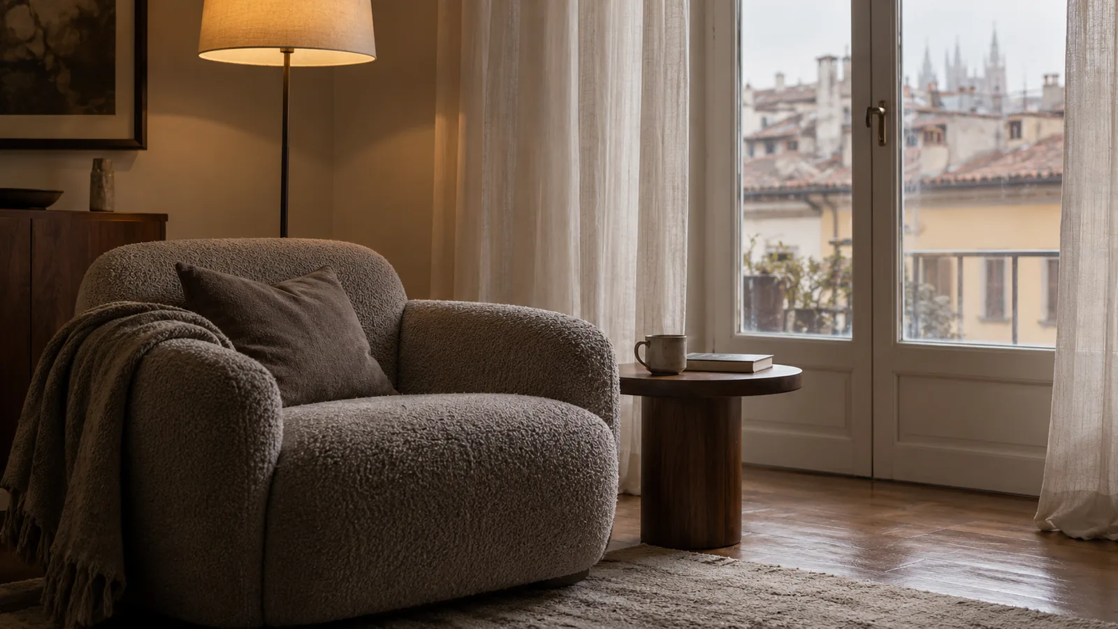 Angolo di un soggiorno milanese con poltrona bouclé, tavolino in noce e finestra con tende leggere